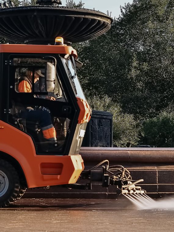 Orange industrial ride-on floor sweeper with water jets cleaning the stone base of an ornate park fountain