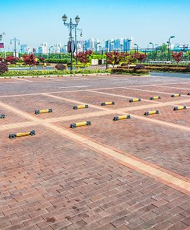 Wide brick-paved outdoor plaza with decorative lampposts, greenery, and a city skyline in the background