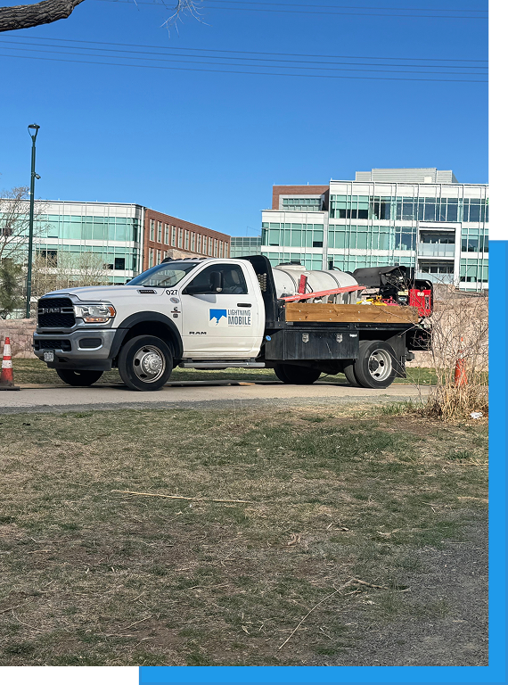White Lightning Mobile RAM flatbed truck with cleaning equipment loaded in the bed, parked near a modern office building campus