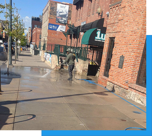 About us 2 Worker pressure washing a wet concrete sidewalk in front of a brick building on a commercial street