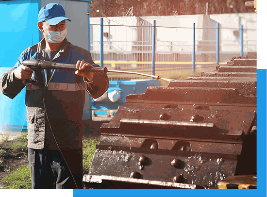 Worker in a cap and coveralls pressure washing rusted industrial steel plates outdoors